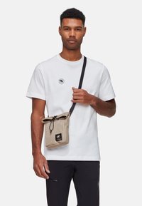 Sac bandoulière en toile beige avec une fermeture à cordon, doté d'un patch logo noir et d'une bandoulière réglable, porté par un mannequin en t-shirt blanc.