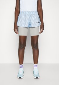 Osoba v tyrkysových vzorovaných sportovních šortkách přes šedé kompresní šortky, v bílých ponožkách s fialovými pruhy a v tyrkysových běžeckých botách.