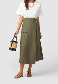 Olijfgroene wikkelrok met een brede tailleband en een decoratieve gesp. Gepaard met een wit t-shirt en een zwarte rieten tas.