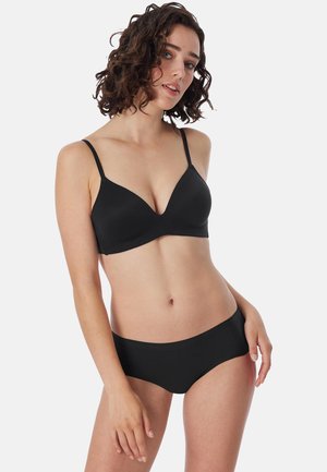 Giovane donna con capelli ricci e scuri, che indossa un reggiseno nero e mutandine nere abbinate, in piedi su uno sfondo bianco.