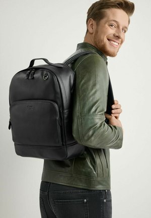 Czarny skórzany plecak o gładkiej fakturze, z zamykanymi na zamek kieszeniami i górnym uchwytem, noszony przez osobę w zielonej skórzanej kurtce.