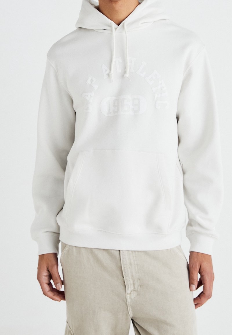Personne portant un sweat à capuche blanc avec une poche avant, portant l'inscription embossée "NAP ATHLETIC 1969" sur le devant, associé à un pantalon beige.
