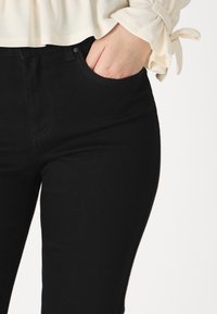 Femme portant un jean noir taille haute, main dans la poche, et un chemisier crème à manches longues avec des poignets noués.