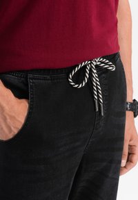 Svarta byxor med en strukturerad yta och elastisk midja, med en flätad svart och vit dragsko. Sidoficka synlig.