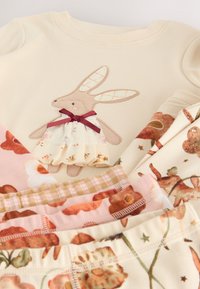 Lys creme top med kaninbroderi og vinrød sløjfe, kombineret med mønstrede blomstrede bukser i pastelrosa og cremefarver. Blødt materiale.