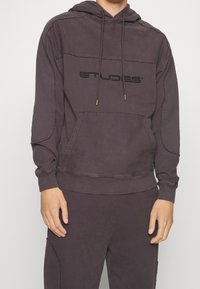 Tmavě fialová mikina s klokankou, šňůrkou a embosovaným logem. Obsahuje ribbenou manžetu a lem a má uvolněný střih.