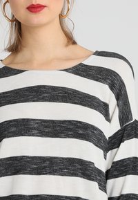 Valkoinen ja musta raidallinen neuletakki, jossa on leveä kaula-aukko ja löysä istuvuus. Kangas näyttää pehmeältä ja siinä on hieno rakenne.