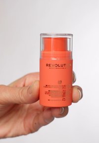Oransje sylinderformet beholder med gjennomsiktig hette, merket "Revolution Makeup." Har en push-dispenser og svart tekst på siden.