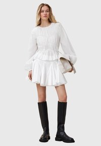 Witte blouse met geplooide textuur, vastgezette taille en uitlopende mouwen, gecombineerd met een uitlopende witte rok en kniehoge zwarte laarzen.