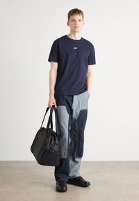 Tricou din bumbac navy cu logo, pantaloni din denim asimetrici în două tonuri, și un rucsac negru. Modelul poartă pantofi negri, stând pe un podea neutră.