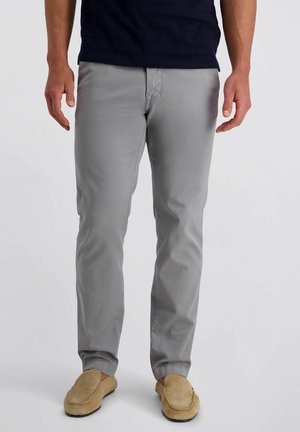 Homme portant un pantalon gris clair à coupe droite et des chaussures à enfiler couleur beige, debout avec les mains détendues le long du corps devant un fond uni.