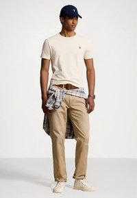 Béžové tričko s malým logem, béžové kalhoty a modrá kostkovaná košile zavázaná v pase. Černá čepice a bílé tenisky doplňují outfit.