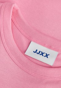 T-shirt de algodão rosa claro com gola redonda canelada. A etiqueta na gola diz "JJXX" em azul sobre um fundo branco. Textura suave.