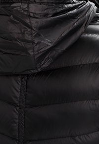 Musta pörröinen takki, jossa on huppu, sileä ja kiiltävä kangas, tilkkutyömuotoilu, jossa on erottuva vaakasauma, sekä säädettävä nyöritys kauluksessa.