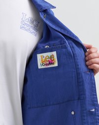 Blue lined shirt features a Crayola label inside, white T-shirt with light blue "Lee Crayola" logo visible. Texture is smooth with visible stitching.