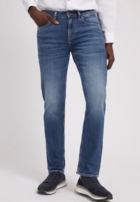 Jeans de denim en un lavado azul medio, con corte ajustado y ligero desgaste. Cuenta con cinco bolsillos, cierre de cremallera y una cinturilla estándar.