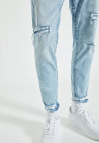 Jean déchiré bleu clair roulé aux chevilles, porté avec des chaussettes blanches et des baskets blanches à lacets sur un fond clair.