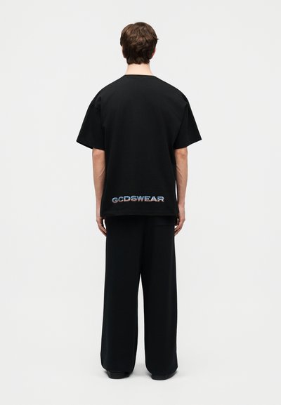Άτομο που φοράει μαύρο υπερμεγέθες μπλουζάκι και παντελόνι με φαρδύ πόδι, στέκεται με το πίσω μέρος προς τα εμπρός, με το κείμενο "GCDSWEAR" κοντά στο τελείωμα.