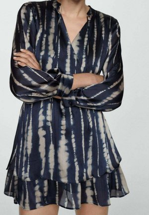 Kobieta w granatowo-beżowej sukience tie-dye z długim rękawem, dekoltem w serek i warstwowym falbaniastym dołem, z rękami skrzyżowanymi na klatce piersiowej.