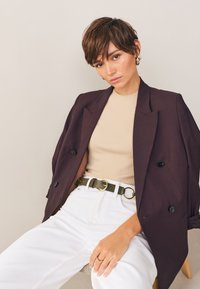 Dwurzędowa bordowa marynarka na dopasowanym beżowym topie, białe jeansy z wysokim stanem oraz oliwkowozielony pasek ze złotą klamrą. Gładka faktura, dopasowany krój.