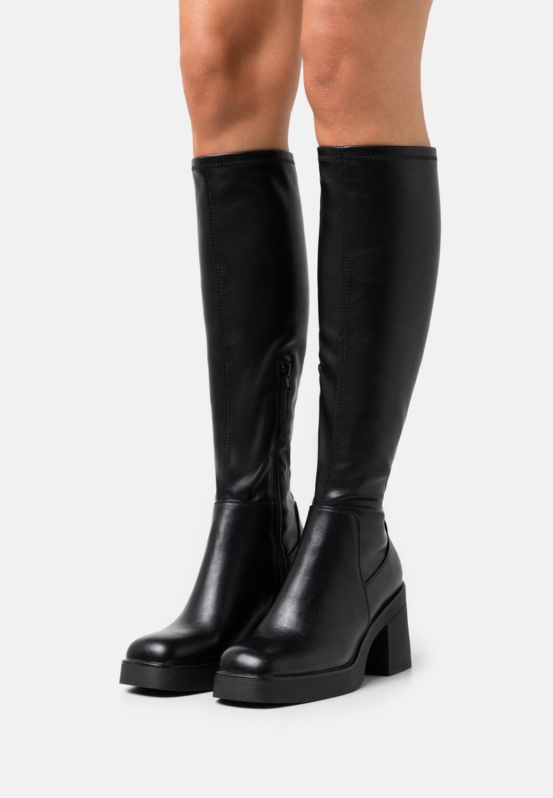 Bottes en cuir noires montant jusqu'aux genoux avec talons blocs et fermetures éclair latérales, portées sur des jambes nues sur fond blanc.