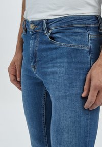 Denimjeans i mellanblå tvätt, med smal passform, subtila blekningar och grå metallknapp. Sidfickor och bältesöglor synliga.