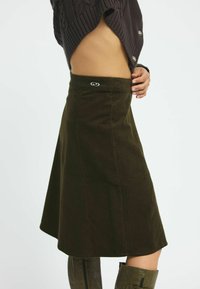Grüne Cord-Rock mit hoher Taille, A-Linie Form und einem bestickten Logo am Taillenbund. Glatte Textur mit sichtbaren Rippungen.