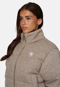 Teksturowana beżowa kurtka puchowa z wysokim kołnierzem, zamkiem błyskawicznym z przodu oraz haftowanym logo na piersi. Miękka tkanina o casualowym stylu.