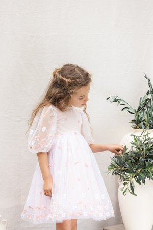 Robe rose clair avec manches courtes bouffantes en tulle transparent. Broderie florale en blanc et pêche sur la jupe en tulle. Un enfant touche une plante en pot.