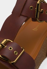 Καφέ δερμάτινο σανδάλι με αγκράφες από μέταλλο σε χρυσό τόνο και φελλό στα τακούνια, με κοντινή εικόνα από τα λουράκια και τη σόλα του ποδιού.