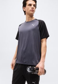 Νέος άνδρας με μαύρη και γκρι αθλητική ενδυμασία Puma κρατώντας ένα διαφανές μαύρο μπουκάλι νερού, κοιτάζοντας προς το πλάι με φόντο μια απλή επιφάνεια.