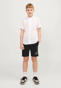 Garçon aux cheveux blonds courts portant une chemise blanche à manches courtes avec boutons, un short noir avec logo, des chaussettes blanches et des baskets noir et blanc, debout.