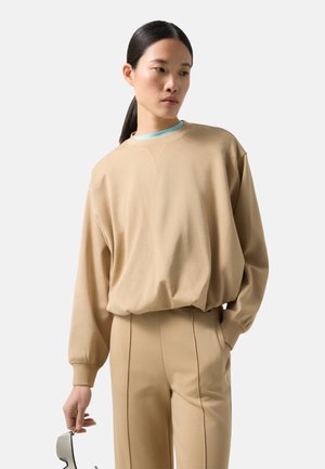 Kvinde med mørkt hår i lav hestehale iført beige sweatshirt og matchende bukser, holder solbriller i højre hånd og kigger til siden.