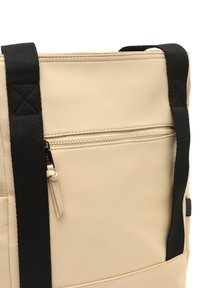 Borsa a spalla beige con manici in tessuto nero, dotata di tasca con zip e una superficie liscia e texturizzata con dettagli di cucitura visibili.