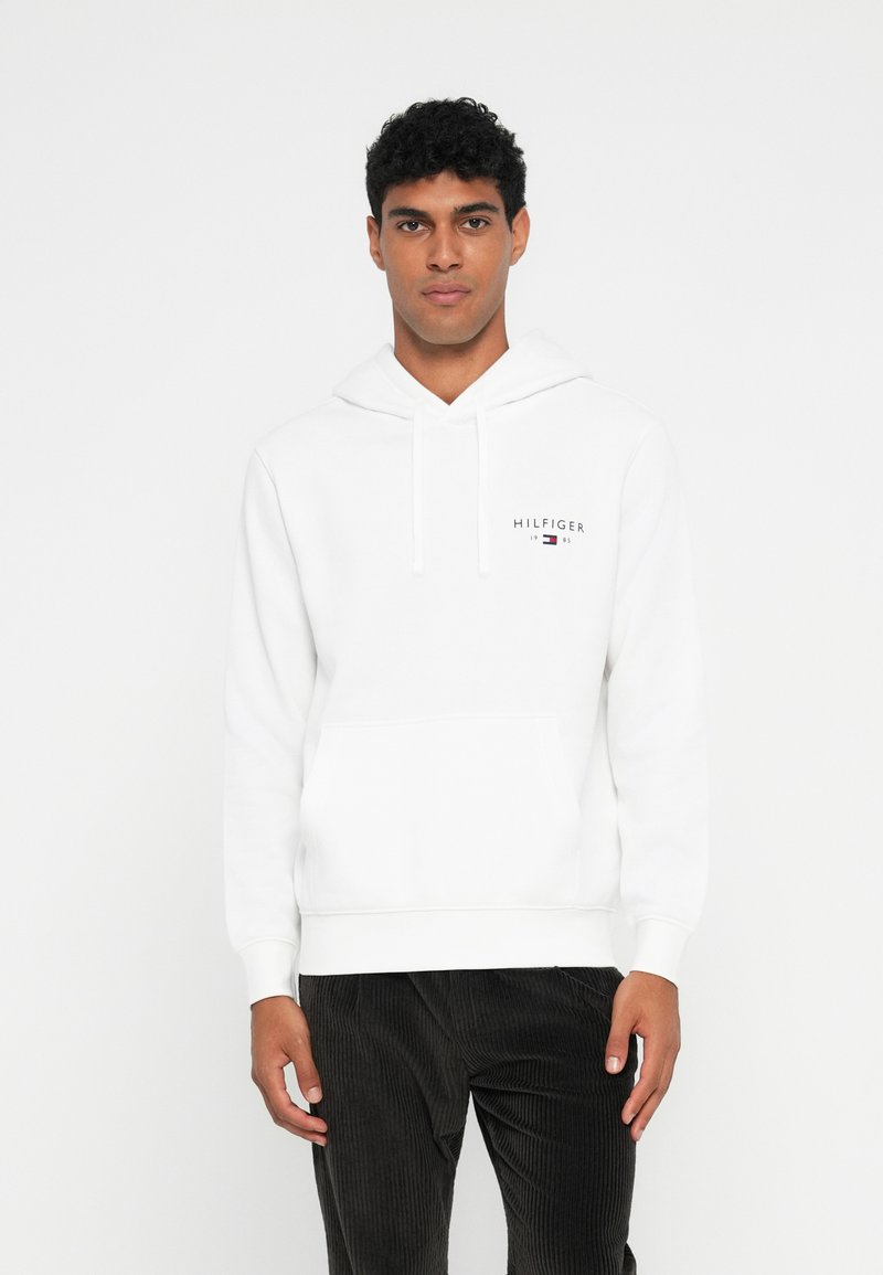 Sweatshirt de algodão branco com bolso frontal, capuz com cordão e logótipo "HILFIGER". Modelo veste calças de veludo canelado pretas.
