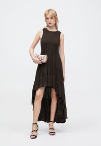 Tamsiai ruda be rankovių suknelė su surinktu bodys, sluoksniuota volanų sijonu, aukštu-žemu kraštu ir modeliu, laikantiu mažą rožinę rankinę. Juodos platforminės basutės.