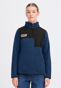 Pullover in pile di colore blu scuro con un pannello frontale nero, design con mezza zip e patch del logo. Presenta una tasca con zip anteriore e una texture morbida.