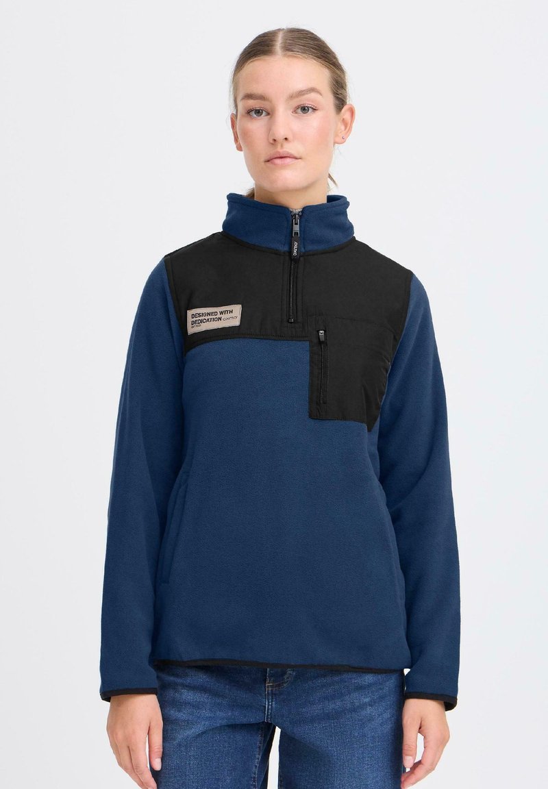 Pullover in pile di colore blu scuro con un pannello frontale nero, design con mezza zip e patch del logo. Presenta una tasca con zip anteriore e una texture morbida.