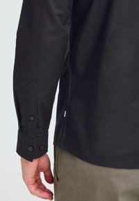 Black long-sleeve shirt with buttoned cuffs and side slits. Fabric appears smooth with a slight sheen. Hand holding shirt at the hem.