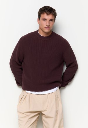 Junger Mann trägt einen dunkelbraunen Strickpullover über einem weißen Hemd mit beigen plissierten Hosen und steht vor einem schlichten weißen Hintergrund.
