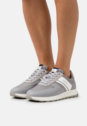 Henkilö, joka käyttää harmaita ja valkoisia JOOP! nauhakenkiä, seisoo valkoisella pinnalla.