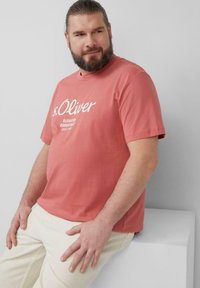 Korallenfarbenes T-Shirt mit kurzen Ärmeln und weißem Text. Verfügt über einen klassischen Rundhalsausschnitt und einen lockeren Schnitt. Kombiniert mit hellen Hosen.