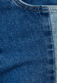 Tessuto denim di colore blu medio con una superficie testurizzata. Presenta cuciture a contrasto lungo il bordo e un pannello di denim più chiaro.
