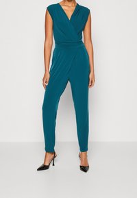 Mujer con un mono sin mangas de color teal con frente cruzado y cintura fruncida, combinado con tacones de slingback negros de punta afilada, de pie frente a una pared blanca.