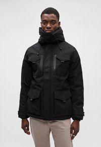 Chaqueta negra aislante con cuello alto, doble cierre, cuatro bolsillos delanteros y cordón ajustable. Textura suave y diseño utilitario.