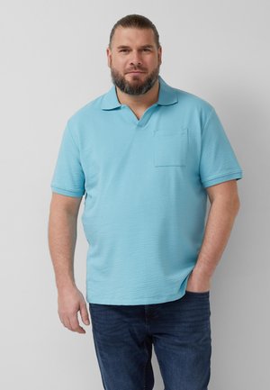 Mann mit Bart, der ein hellblaues kurzärmeliges Poloshirt und dunkle Jeans trägt, steht mit einer Hand in der Tasche vor grauem Hintergrund.