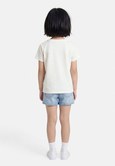 Παιδί που στέκεται στραμμένο προς τα πίσω, φοράει απλό λευκό μπλουζάκι, ανοιχτό μπλε τζιν σορτς, λευκές κάλτσες και λευκά αθλητικά παπούτσια.