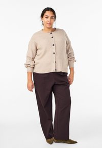 Béžový rebrovaný sveter na gombíky s okrúhlym výstrihom, zapínaný vpredu, s širokými rukávmi, v kombinácii s tmavohnedými širokými nohavicami a zelenými špicatými topánkami.