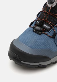 Chaussure de randonnée bleue et noire avec un dessus texturé, un renfort en caoutchouc à l'avant et un système de laçage élastique. Présente le logo GORE-TEX et une semelle robuste.