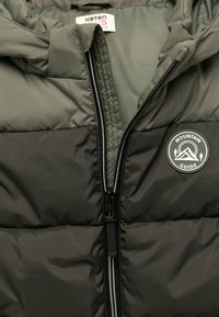 Chaqueta acolchada verde oscuro con cremallera frontal. Presenta un parche redondo con el logo "Mountain Guide" y textura acolchada.